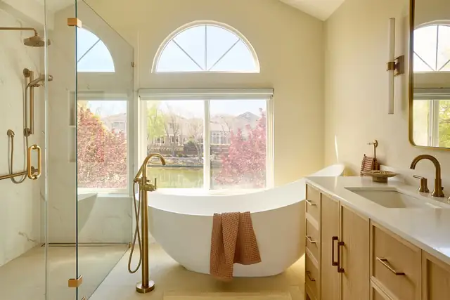 Alameda primary bathroom with a freestanding tub under an arched window, glass shower and wood vanity with brass fixtures