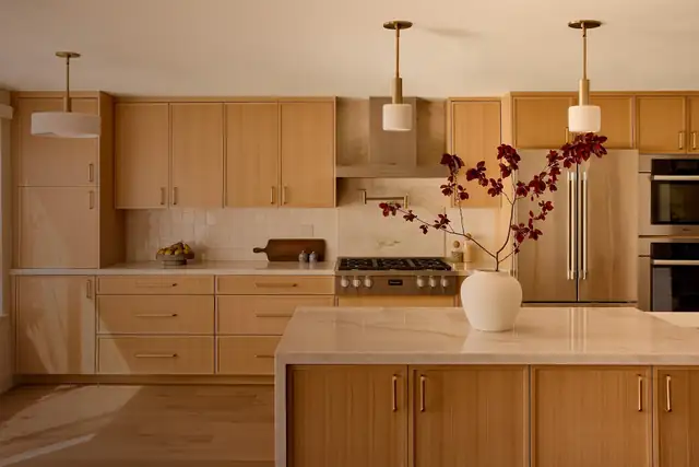 Warm white oak kitchen with flat-panel cabinets, brass pendant lights, a stone-top island and built-in wall ovens